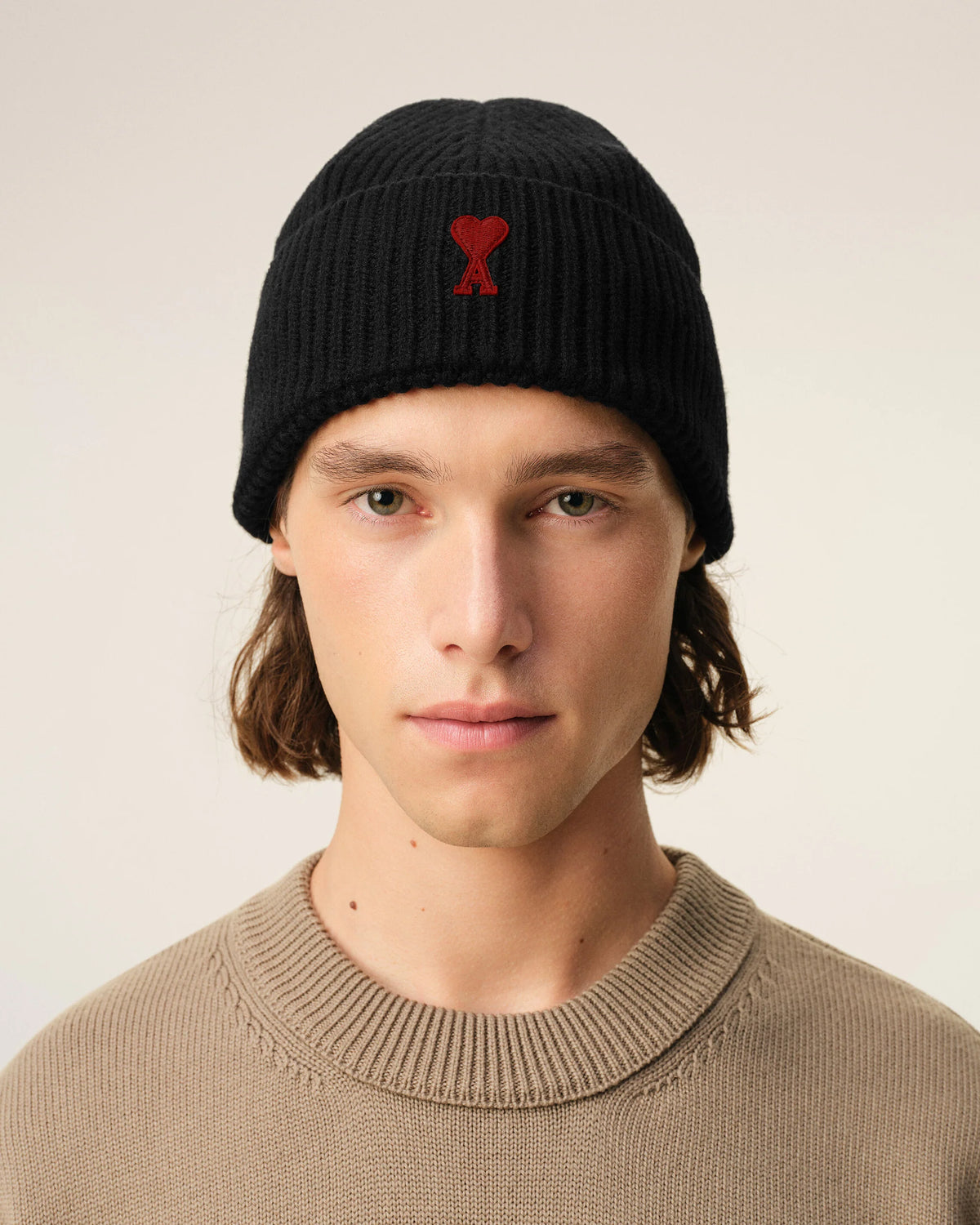 A black ribbed knit beanie hat featuring a small red embroidered heart logo with a letter A on the front cuff, set against a plain off-white background.