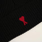 A black ribbed knit beanie hat featuring a small red embroidered heart logo with a letter A on the front cuff, set against a plain off-white background.