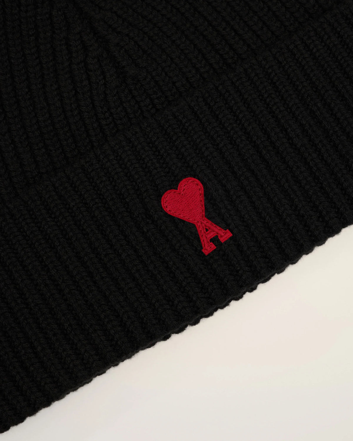 A black ribbed knit beanie hat featuring a small red embroidered heart logo with a letter A on the front cuff, set against a plain off-white background.
