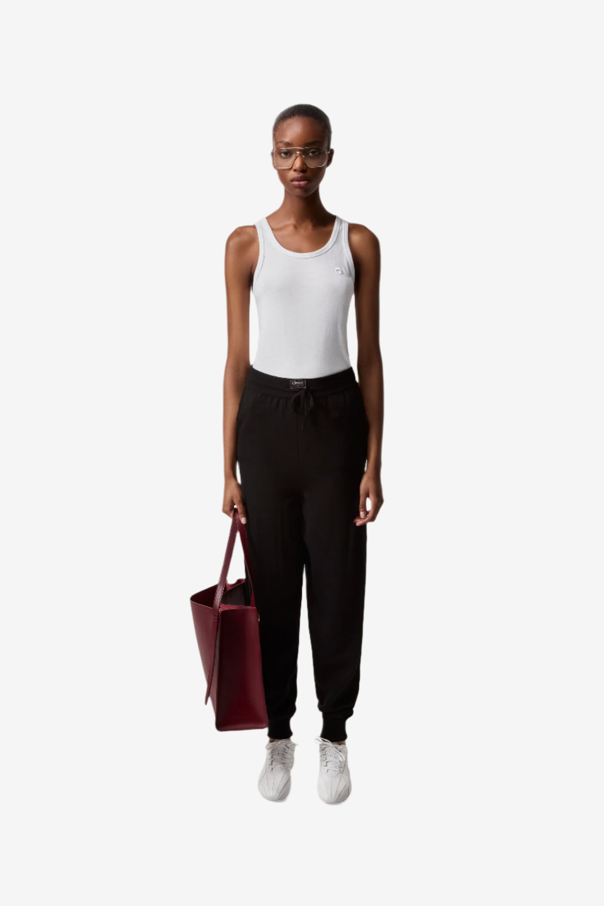Person wearing a white tank top, black pants, and white sneakers, holding a red bag on a white background