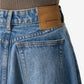 Close-up of blue jeans with a brown leather patch displaying 'ami alexandre mattiussi' on a neutral background.