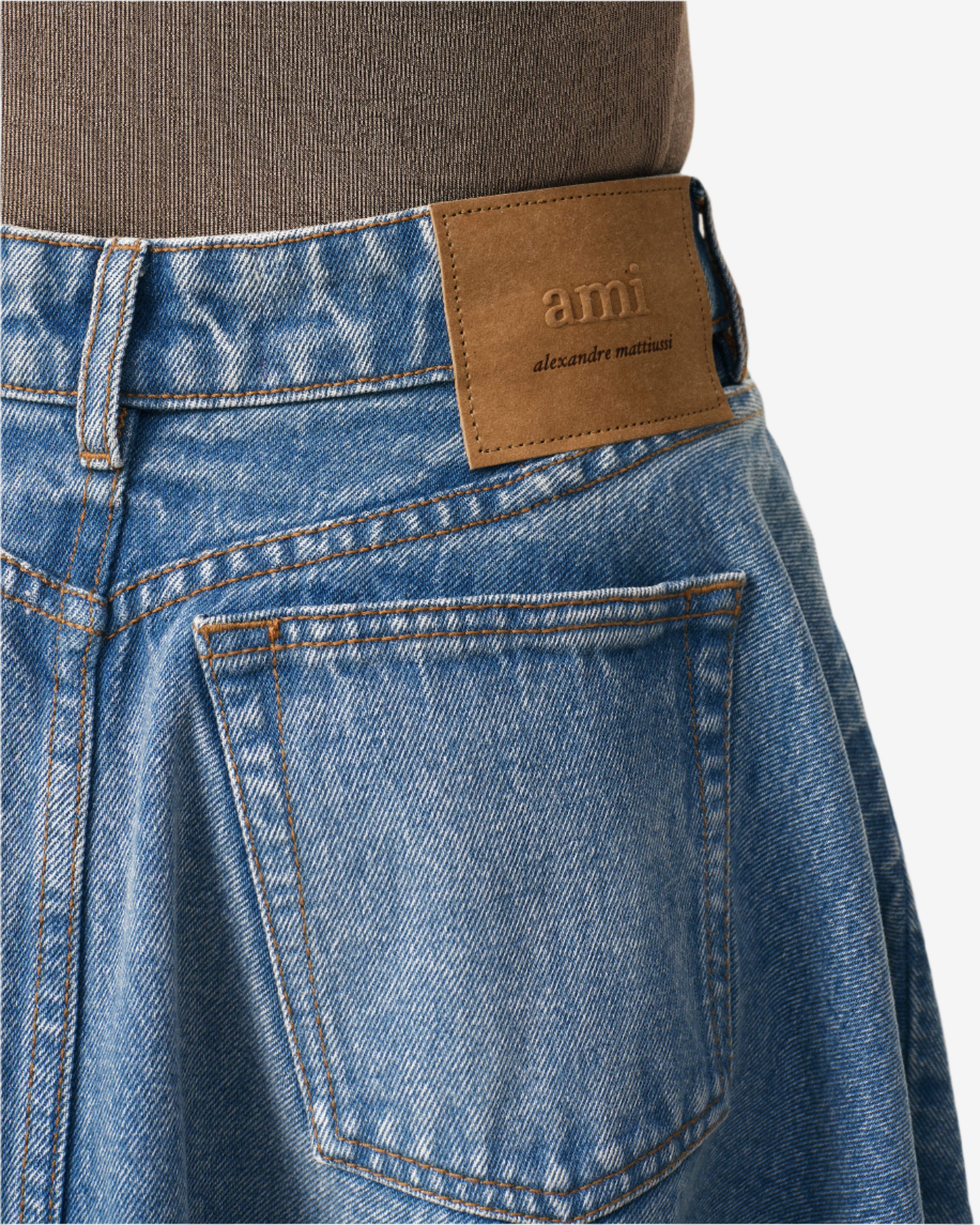 Close-up of blue jeans with a brown leather patch displaying 'ami alexandre mattiussi' on a neutral background.