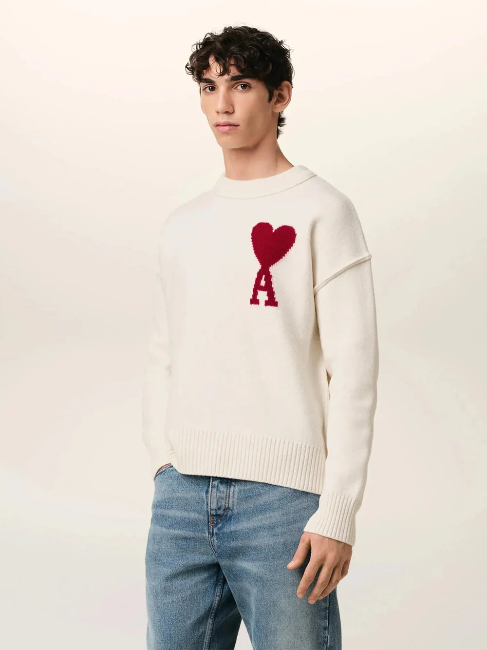 Person wearing a white sweater with a red heart and letter 'A' design, paired with blue jeans on a light background.