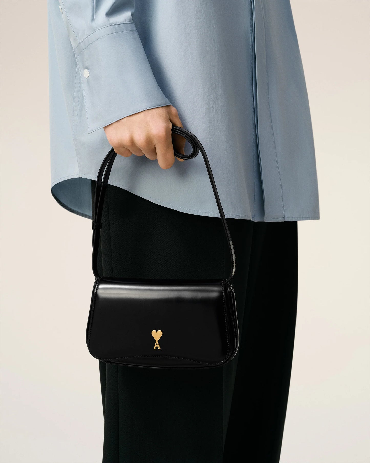 Person holding a black handbag with a gold emblem against a neutral background ami paris