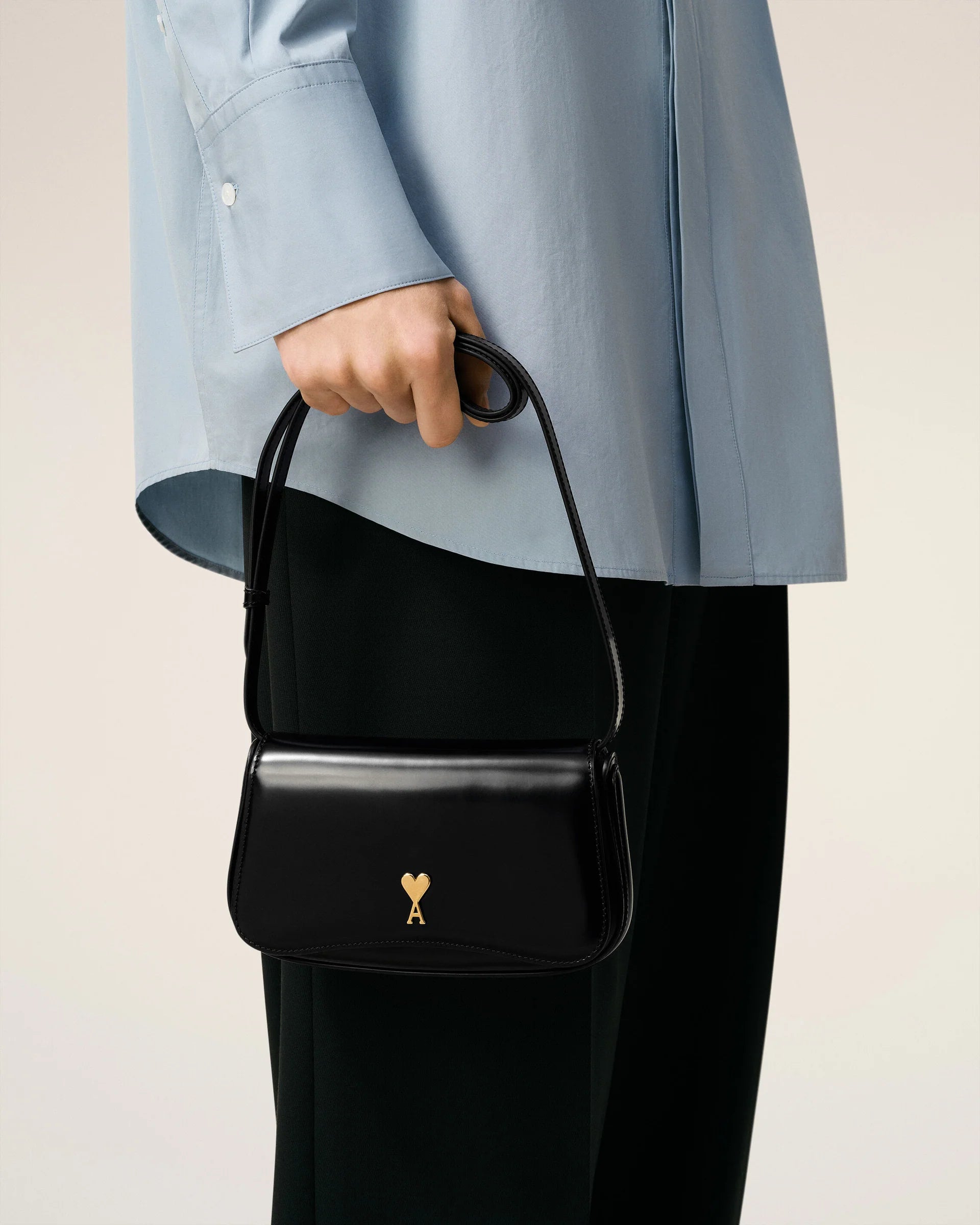Person holding a black handbag with a gold emblem against a neutral background ami paris