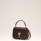 Burgundy handbag with a gold heart and letter 'A' emblem on a beige background