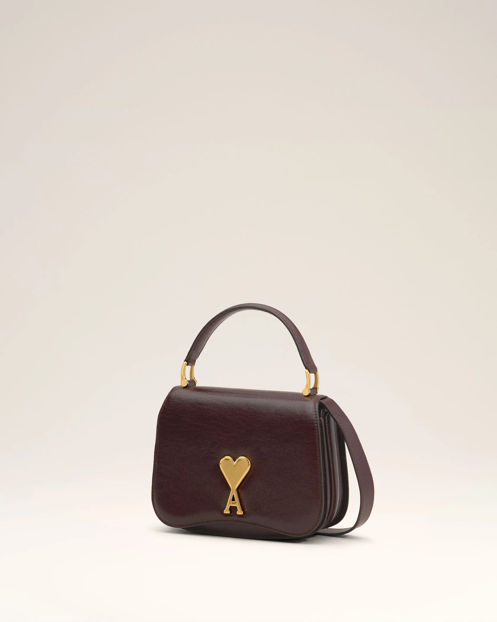 Burgundy handbag with a gold heart and letter 'A' emblem on a beige background