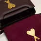Burgundy handbag with a gold heart and letter 'A' emblem on a beige background