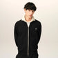 A black zip-up jacket with cream ribbed collar, cuffs, and hem, featuring a small embroidered logo on the left chest, displayed on a mannequin against a light beige background.