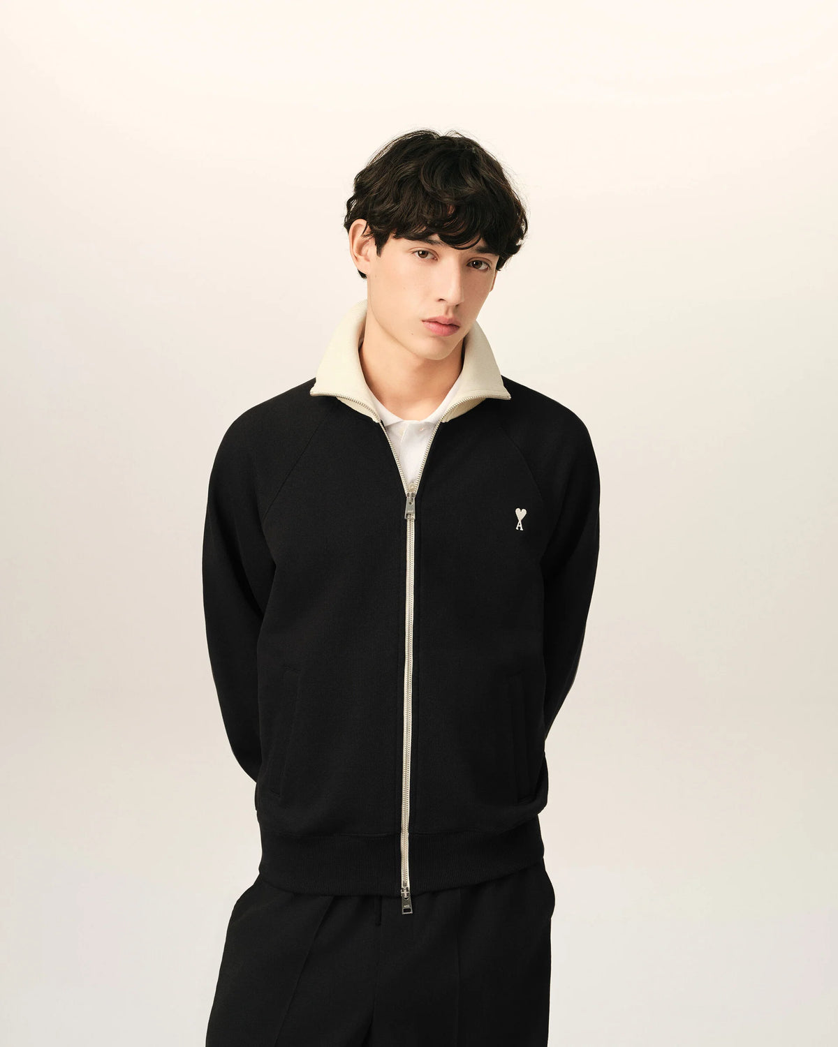 A black zip-up jacket with cream ribbed collar, cuffs, and hem, featuring a small embroidered logo on the left chest, displayed on a mannequin against a light beige background.