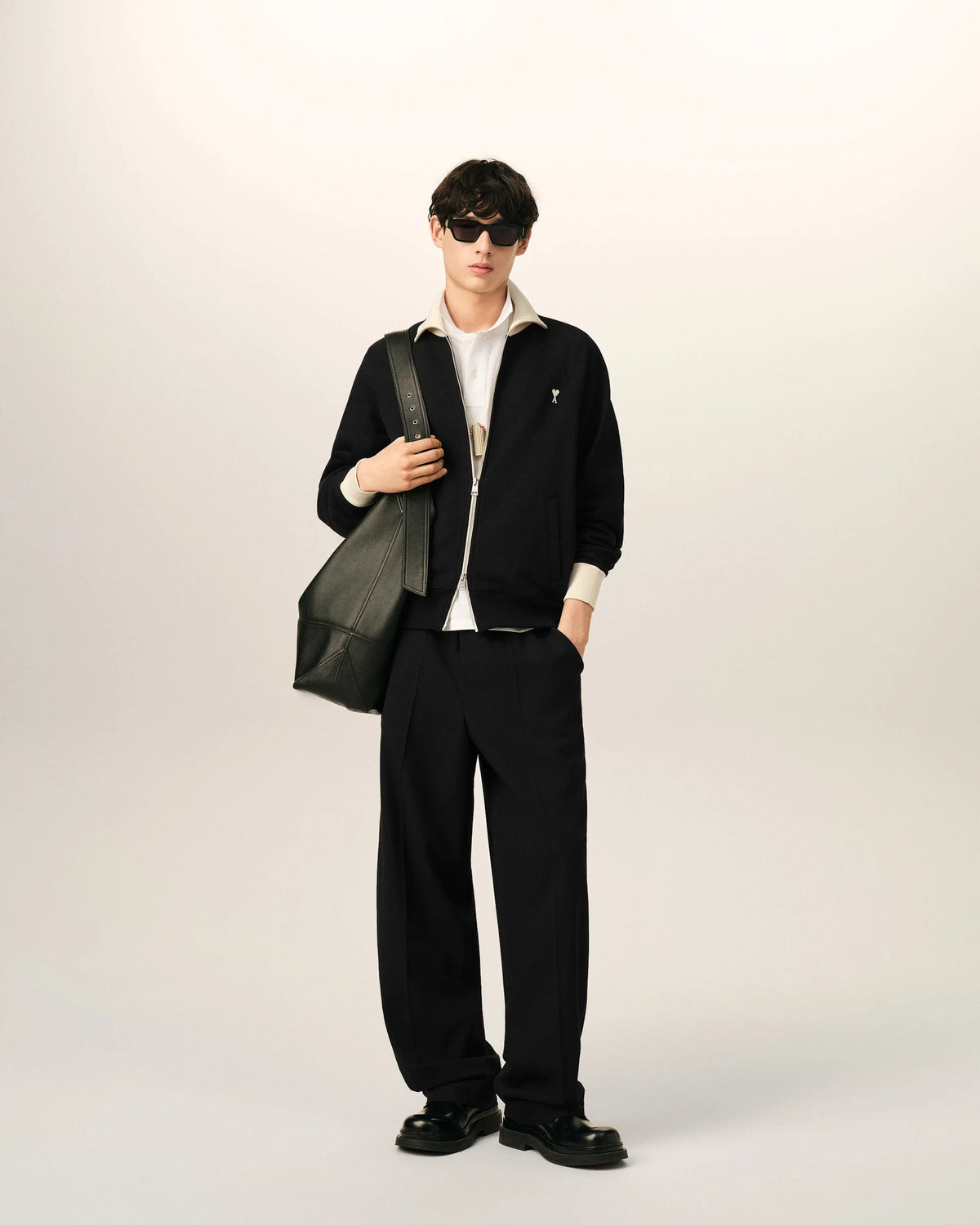 A black zip-up jacket with cream ribbed collar, cuffs, and hem, featuring a small embroidered logo on the left chest, displayed on a mannequin against a light beige background.
