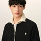 A black zip-up jacket with cream ribbed collar, cuffs, and hem, featuring a small embroidered logo on the left chest, displayed on a mannequin against a light beige background.