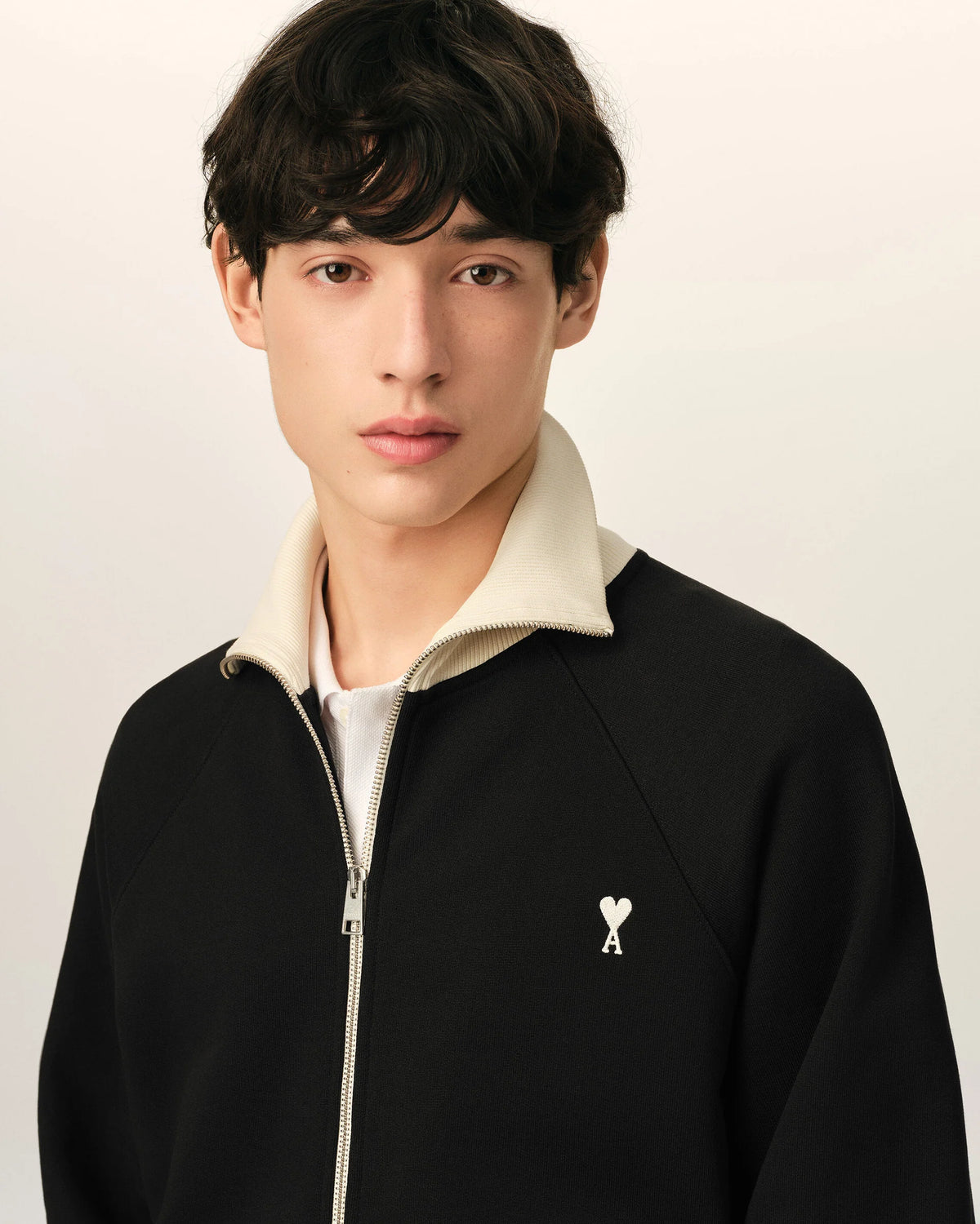 A black zip-up jacket with cream ribbed collar, cuffs, and hem, featuring a small embroidered logo on the left chest, displayed on a mannequin against a light beige background.