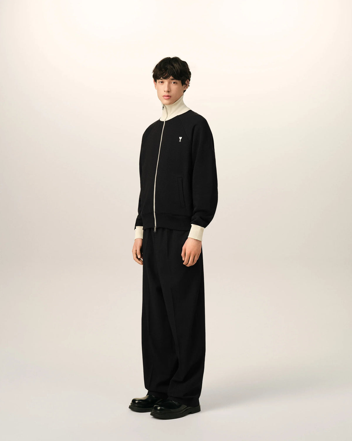 A black zip-up jacket with cream ribbed collar, cuffs, and hem, featuring a small embroidered logo on the left chest, displayed on a mannequin against a light beige background.