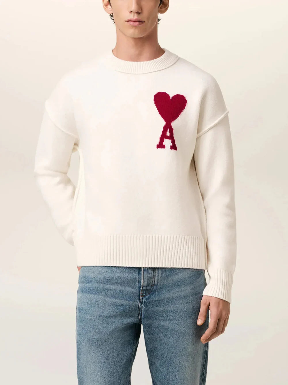 Person wearing a white sweater with a red heart and letter 'A' design, paired with blue jeans on a light background.
